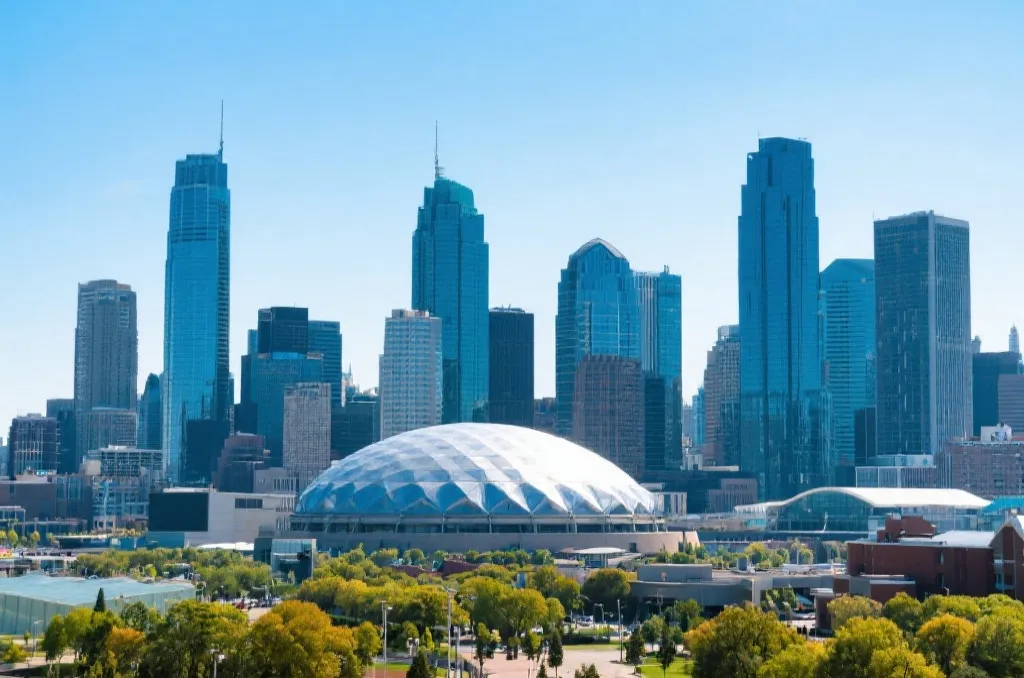 A modern North American city skyline with skyscrapers and a generic stadium dome under a sunny sky.